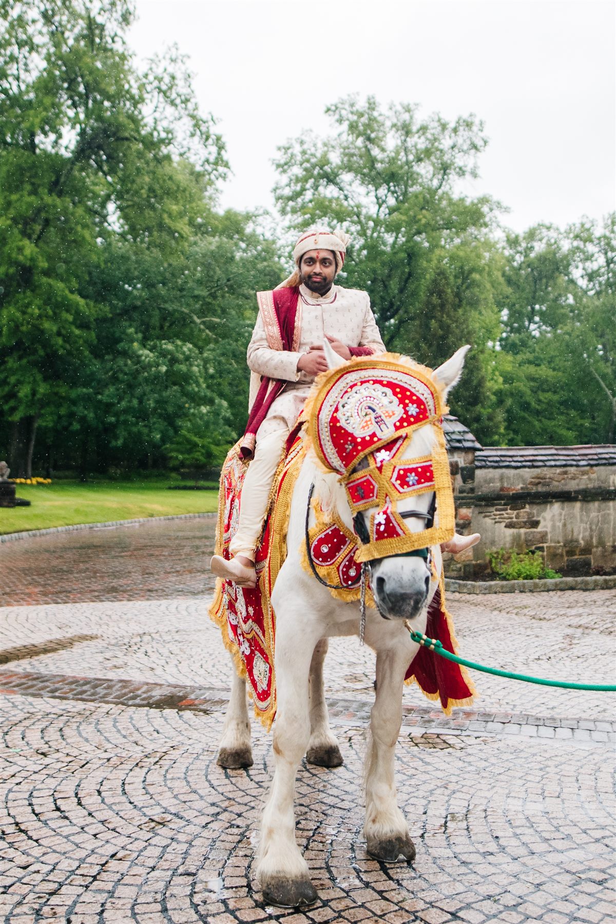 The baraat arrives, the groom on a white horse on the cobblestone drive